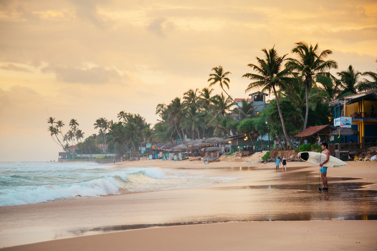 Surfer stands on the sand at Narigama beach in Hikkaduwa during sunset, with a board under his arm.